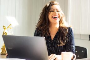Woman smiling online shopping with laptop at desk in sunlight