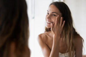 Smiling girl in bath towel looking in mirror, applying moisturizing facial cream